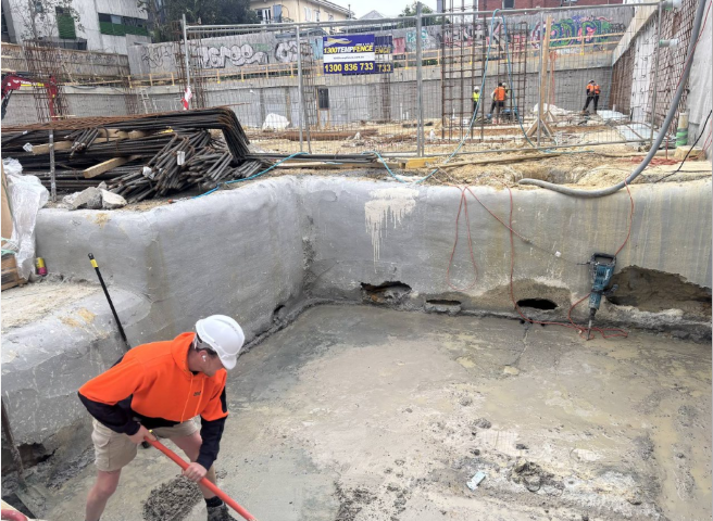 Hulk worker in a white branded hard hat and orange high-visibility hoodie working in a concrete foundation pit on a closely managed site