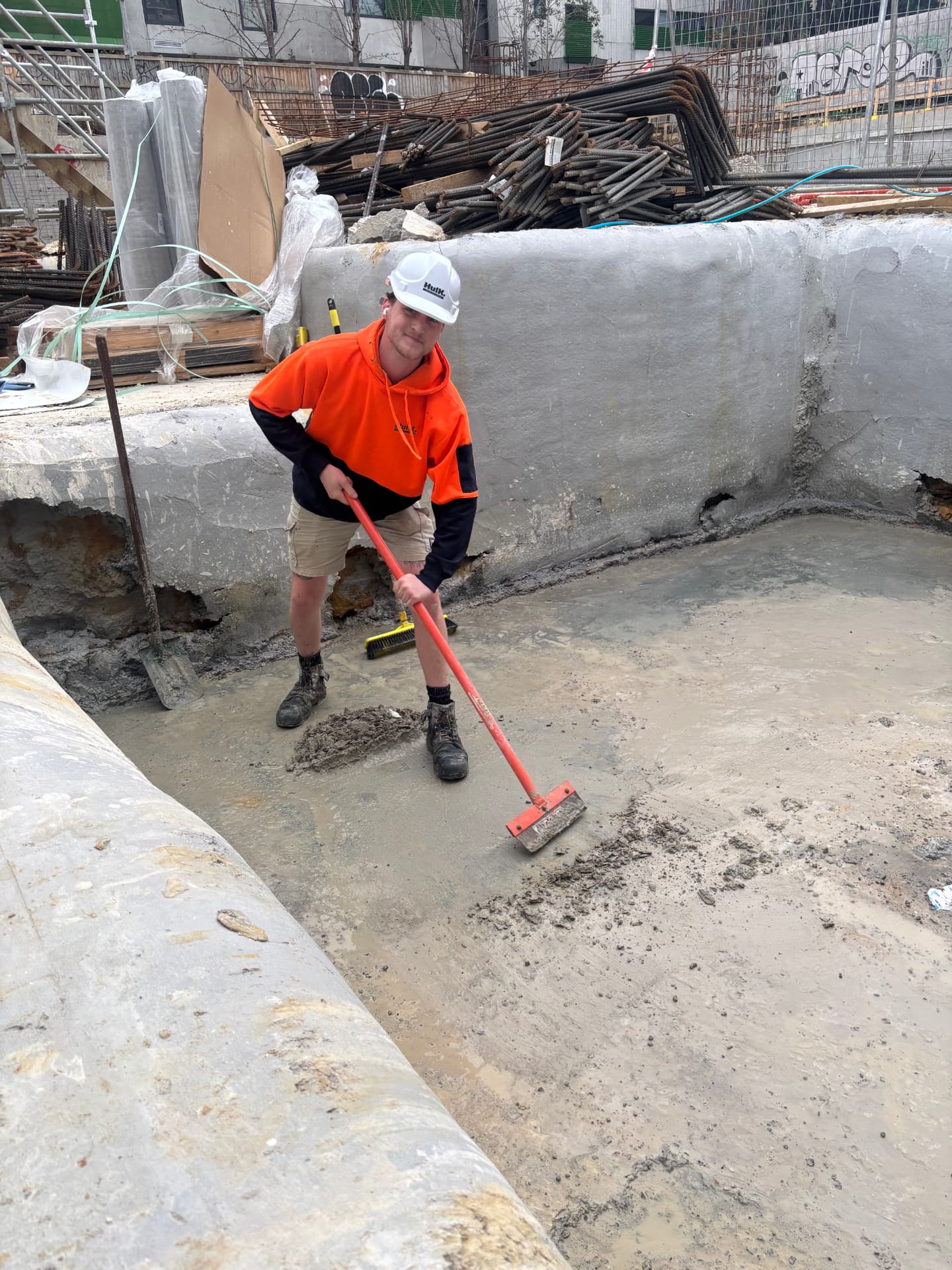Hulk worker in a white hard hat and orange hoodie squeegeeing a freshly poured concrete slab inside a foundation pit