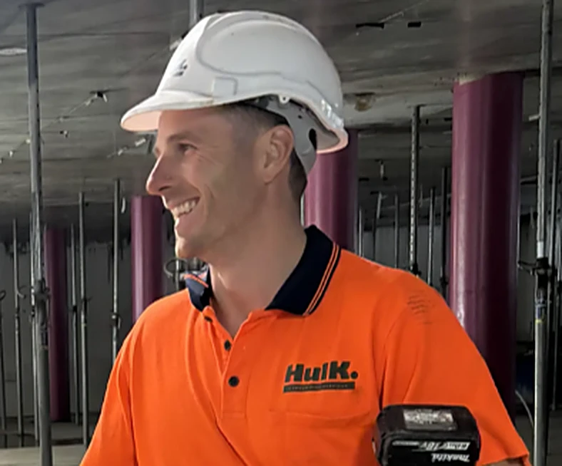 Hulk carpenter in a white branded hard hat and orange Hulk polo smiling on an indoor commercial site